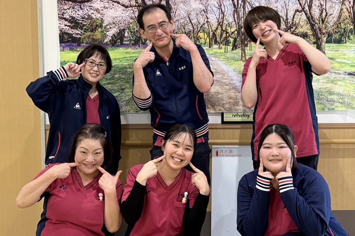 施設スタッフが桜並木を背景に笑顔でポーズを取っている記念写真