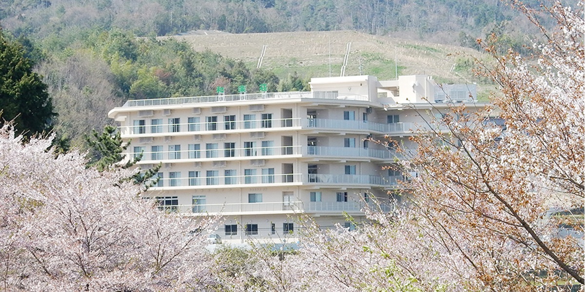 桜並木に囲まれた介護施設の建物外観。春の季節感が表現されている。